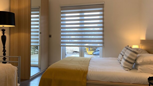 A bedroom with a bed, a bedside lamp, a mirror, and a window with blinds. - Roath Retreat (Cardiff)