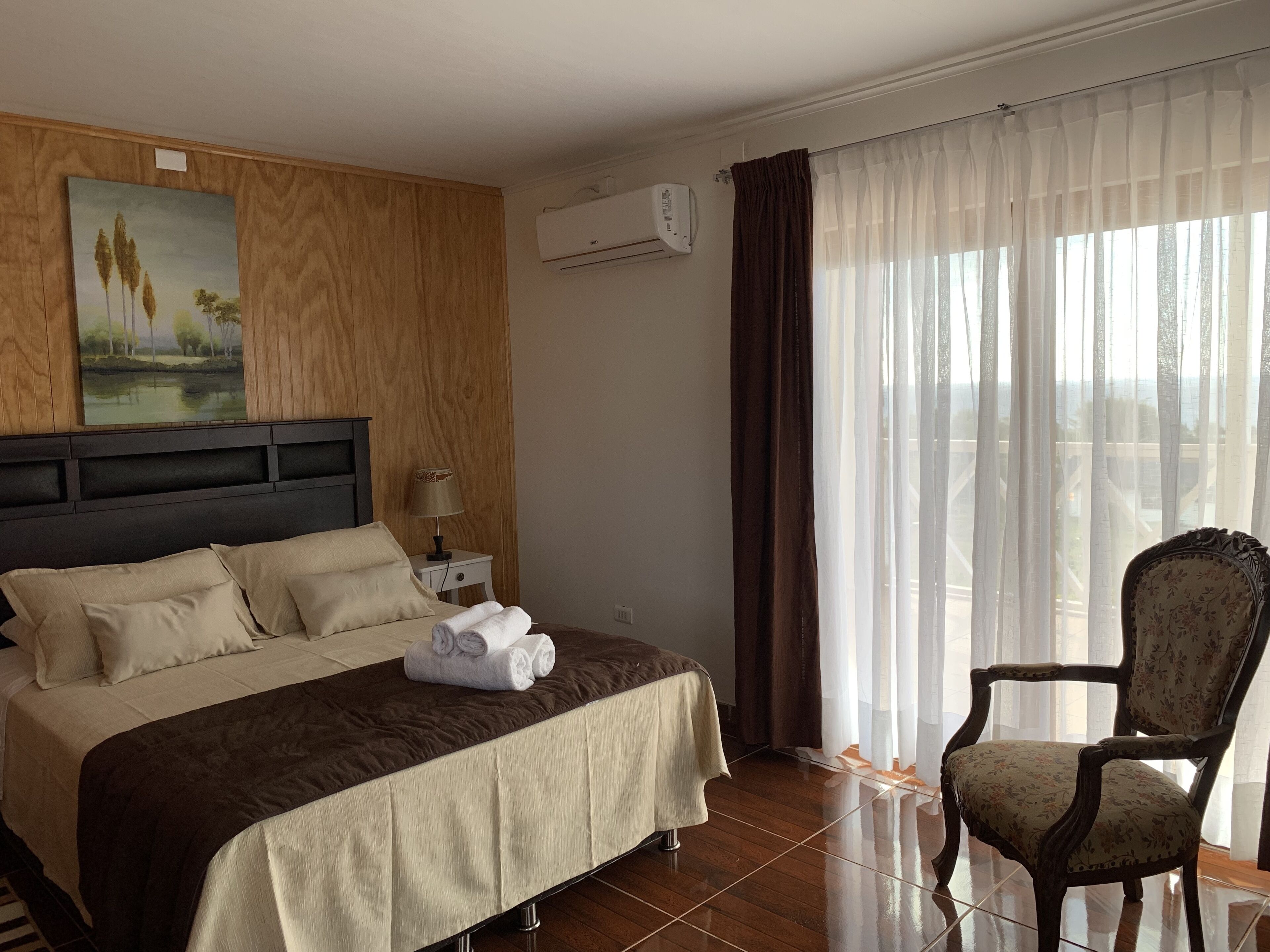 Een hotelkamer met een bed, een stoel, een schilderij, een airconditioner en een raam met gordijnen.