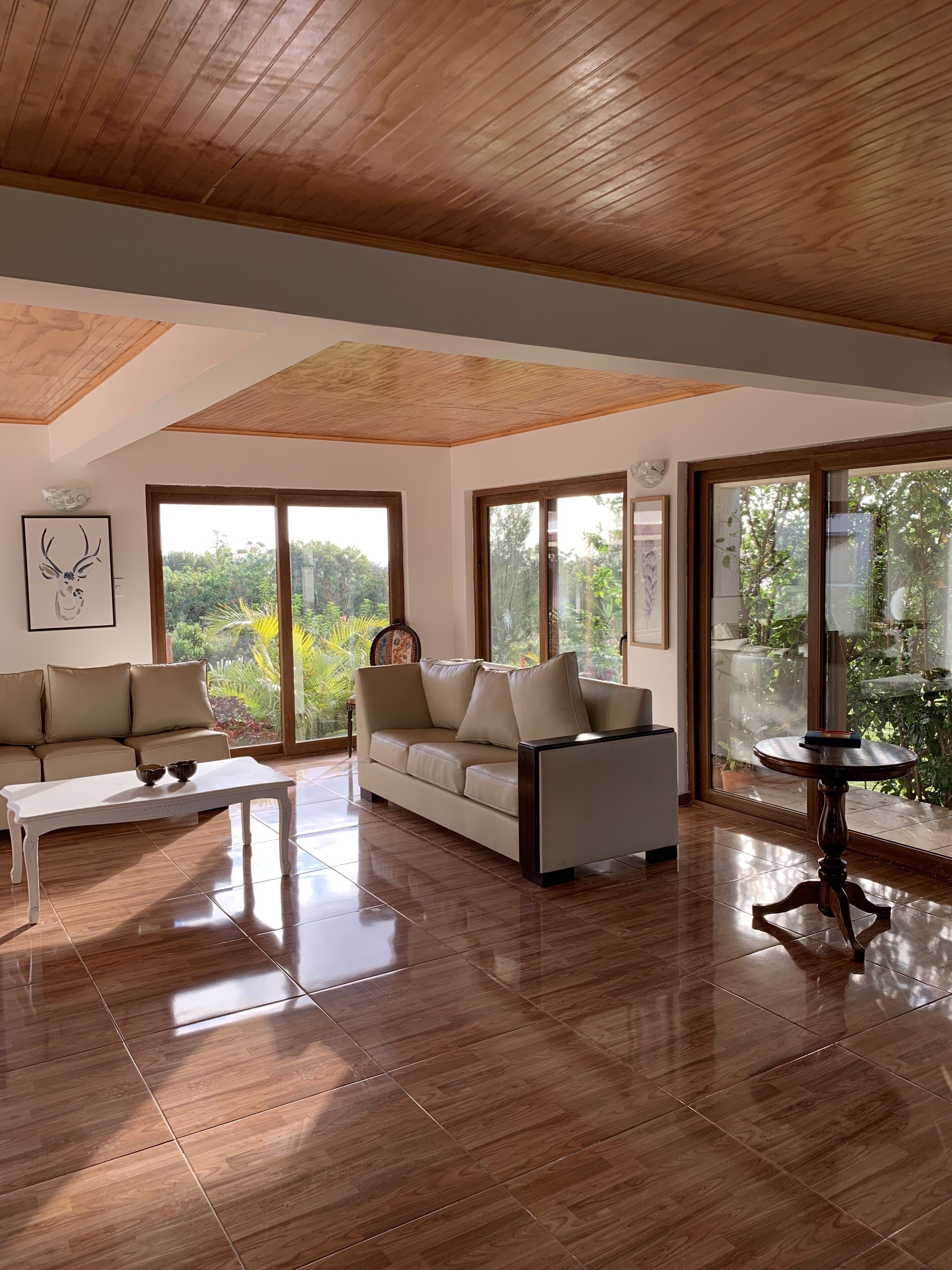 A spacious living room with a wooden ceiling, large windows, a beige sofa, a white coffee table, and a wooden side table.