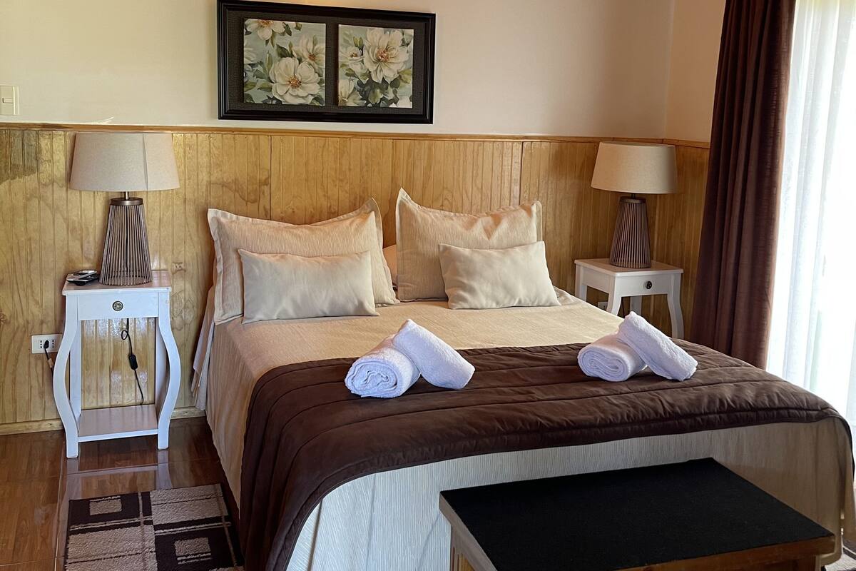 Hotelkamer met een houten plafond, een groot bed met een bruine plaid, twee nachtkastjes met lampen, een wandlamp en een lijst met bloemkunstwerk.