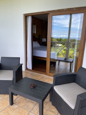 A balcony with wicker furniture, a bed with a wooden headboard, and a view of greenery and the ocean. - Hotel Teanehi (Hanga Roa)
