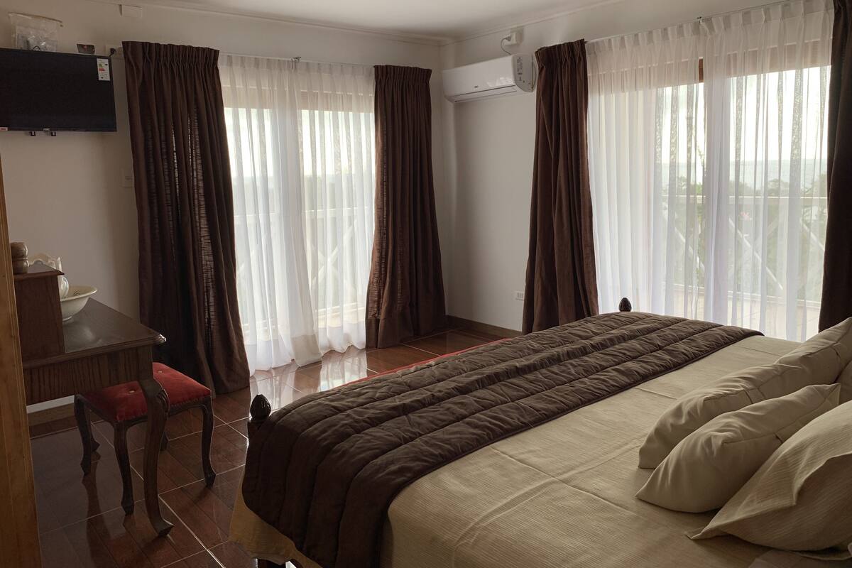 Een hotelkamer met een bed, een bureau en een stoel. Gordijnen zijn aanwezig bij de ramen, en een airconditioningunit is aan de muur bevestigd.