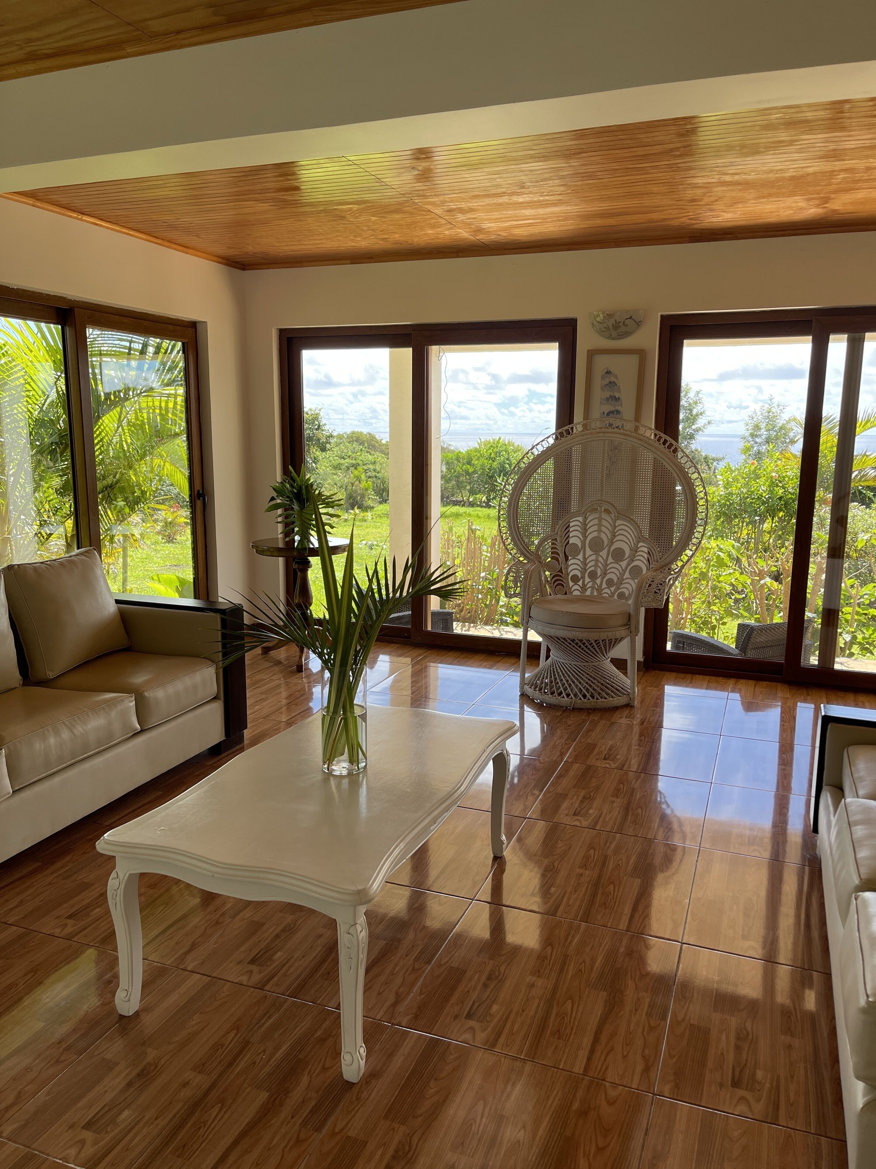 A spacious living room with a beige sofa, a white coffee table, and a wicker chair. Large windows offer views of greenery and the sea.