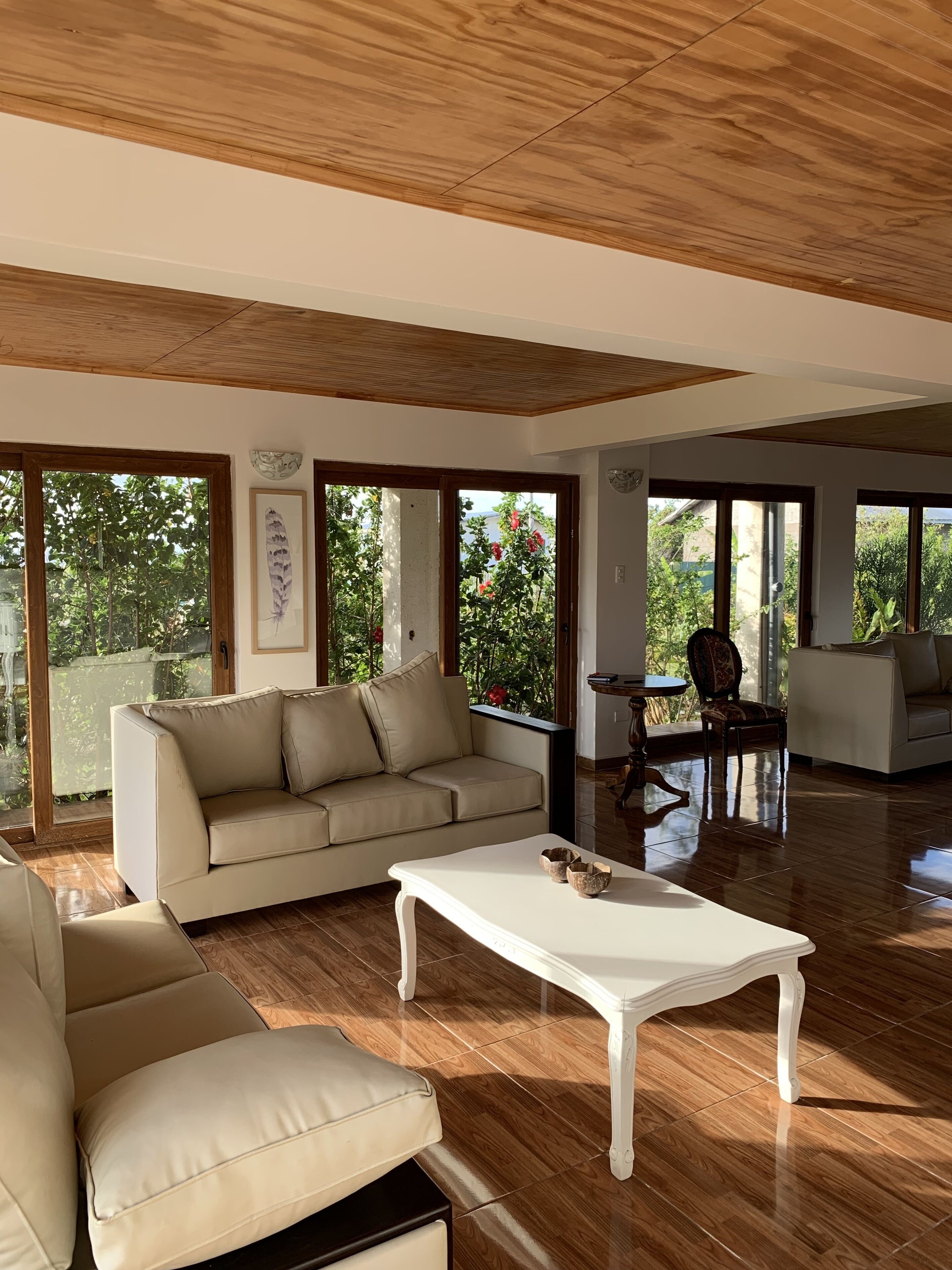 A spacious living room with a beige sofa, a white coffee table, and a dining area with a table and chairs. Large windows provide views of greenery outside.
