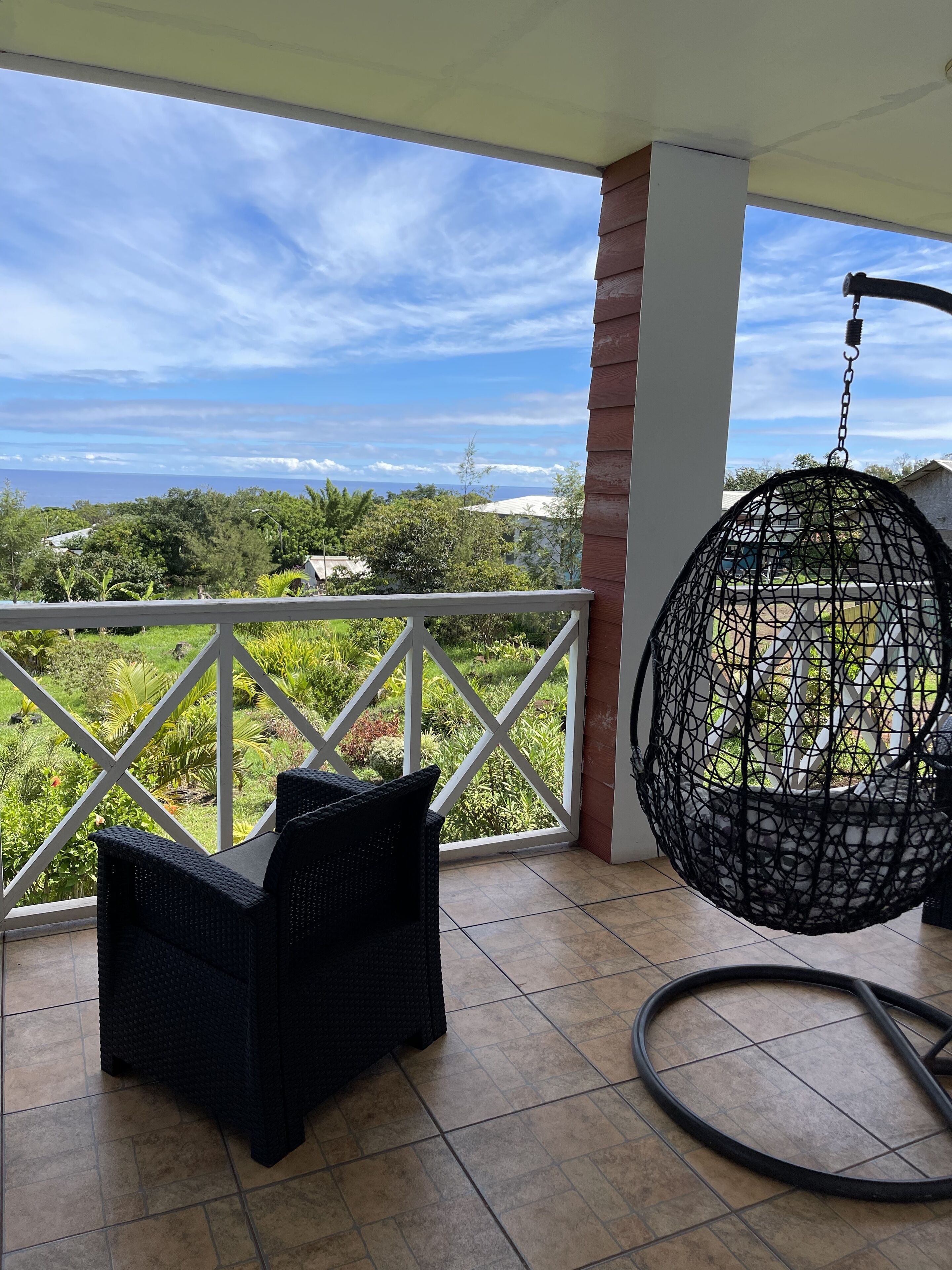 A balcony with a hanging chair and a table, overlooking a lush garden and a distant view of the ocean.