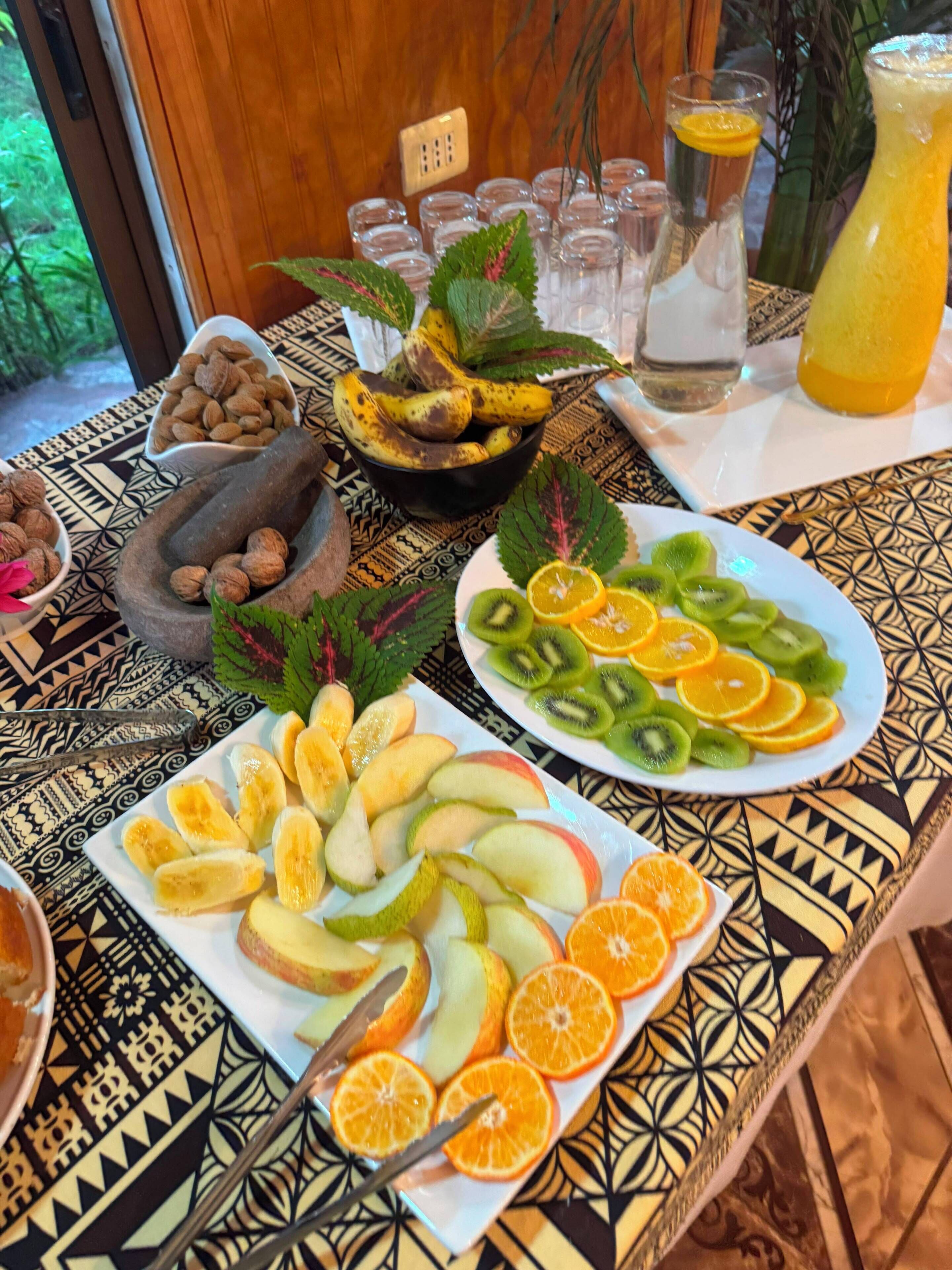 Una mesa con frutas y frutos secos, una jarra de vidrio y un mantel estampado.