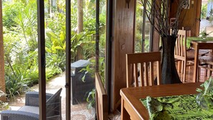 A wooden interior with large sliding glass doors leading to a garden. There are wicker furniture and a wooden table with a green patterned tablecloth. - Hotel Teanehi (Hanga Roa)