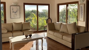 A spacious living room with a wooden ceiling, large windows offering a view of greenery, a beige sofa, and a coffee table. - Hotel Teanehi (Hanga Roa)