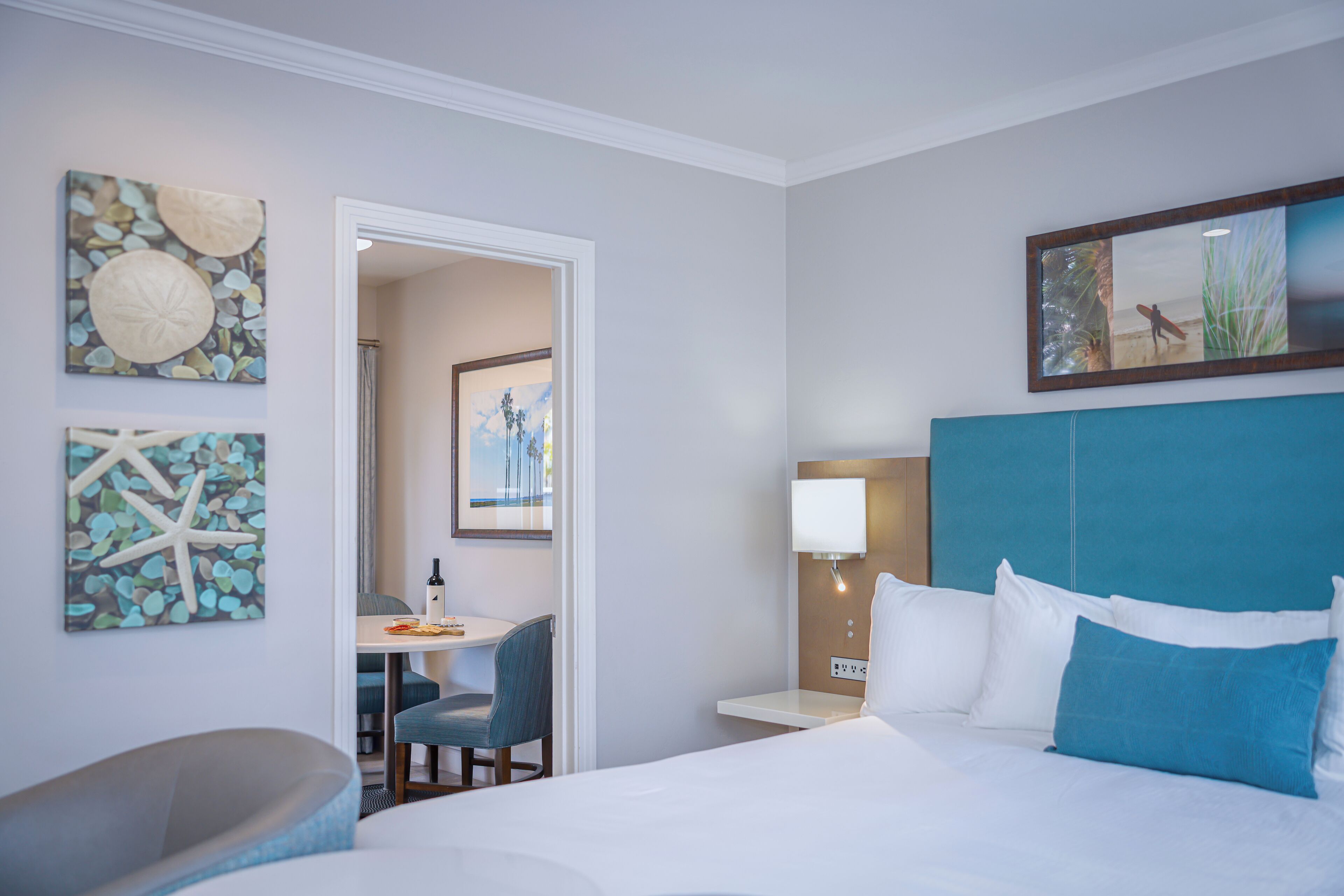 A hotel room with a bed, a desk, and a chair. There is a painting on the wall and a picture above the bed.