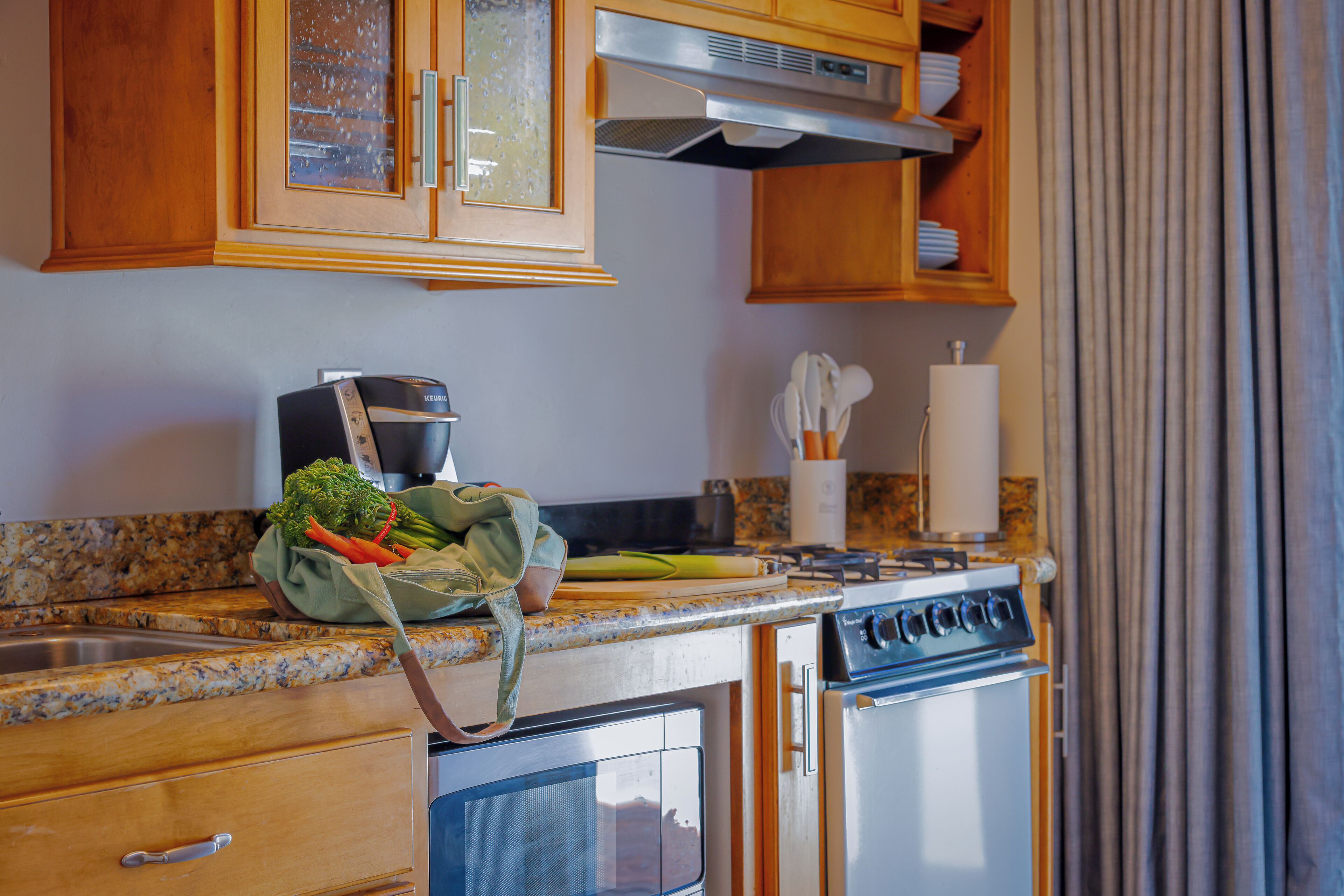 Une cuisine avec des meubles de rangement en bois, une cuisinière, un four, un évier et un plan de travail sur lequel se trouve un sac de légumes.