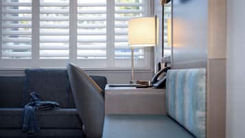A modern hotel room with a sofa, a bedside table with a lamp, and a window with blinds.