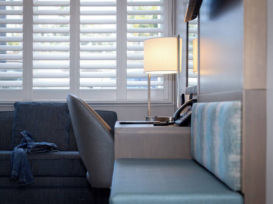 A modern hotel room with a sofa, a bedside table with a lamp, and a window with blinds.