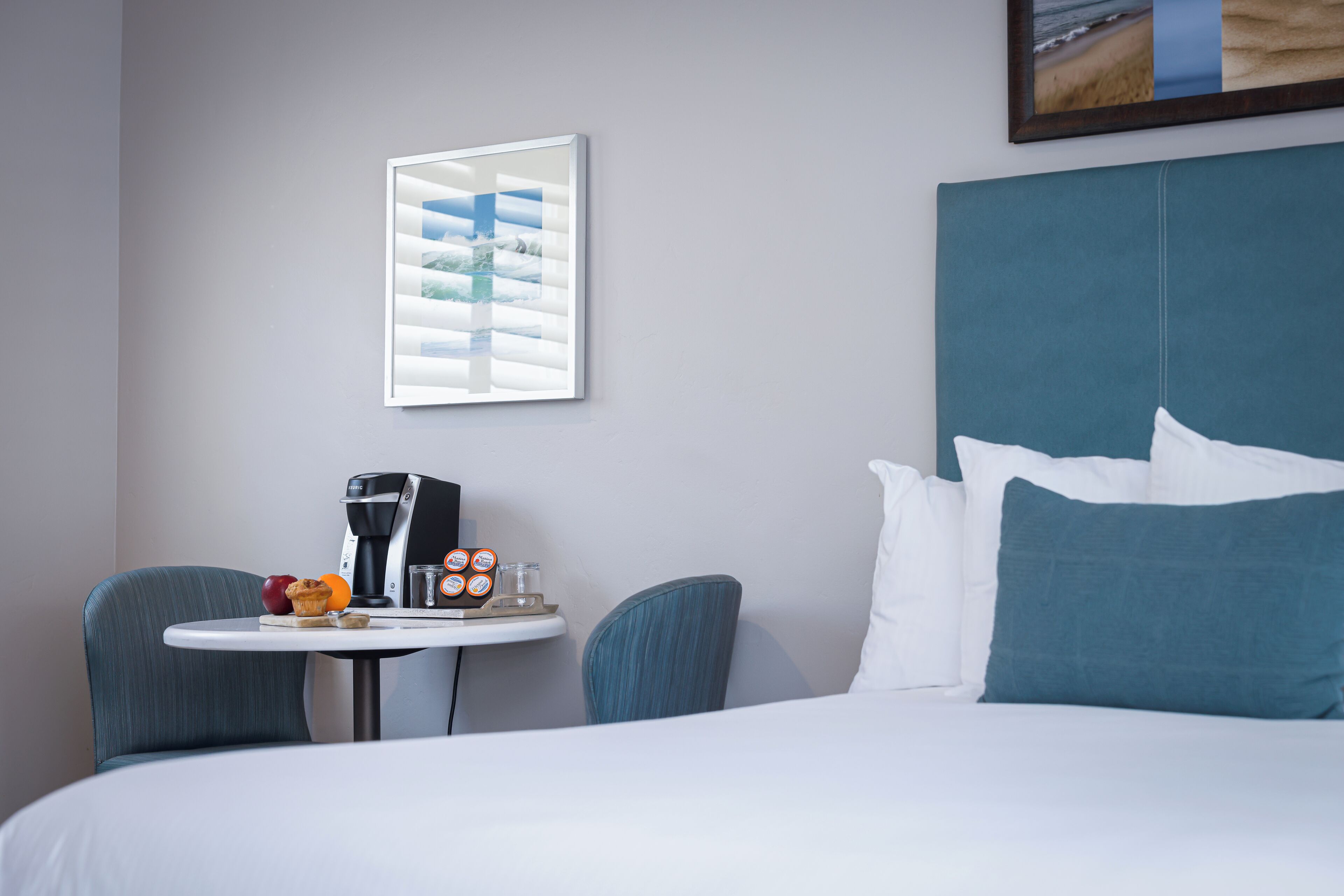 Una habitación de hotel con una cama, una mesita con una cafetera, un muffin y un vaso de agua.