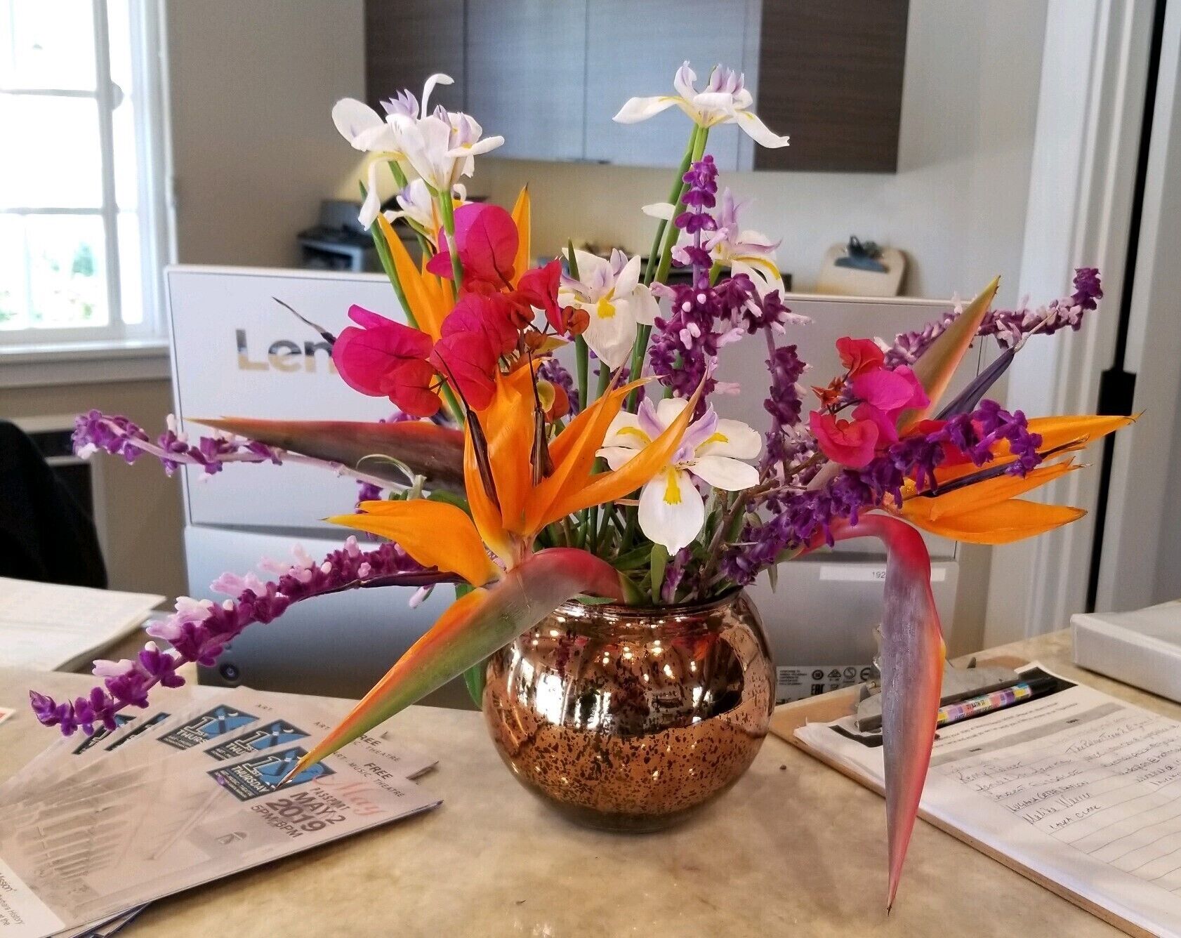 Un arrangement floral dans un vase en cuivre, posé sur une table avec des papiers et un ordinateur portable en arrière-plan.