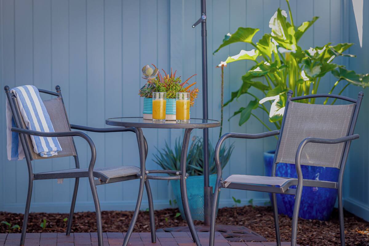 Two metal chairs with blue and white striped towels, a small round table with two glasses of orange juice, and a potted plant in the background.