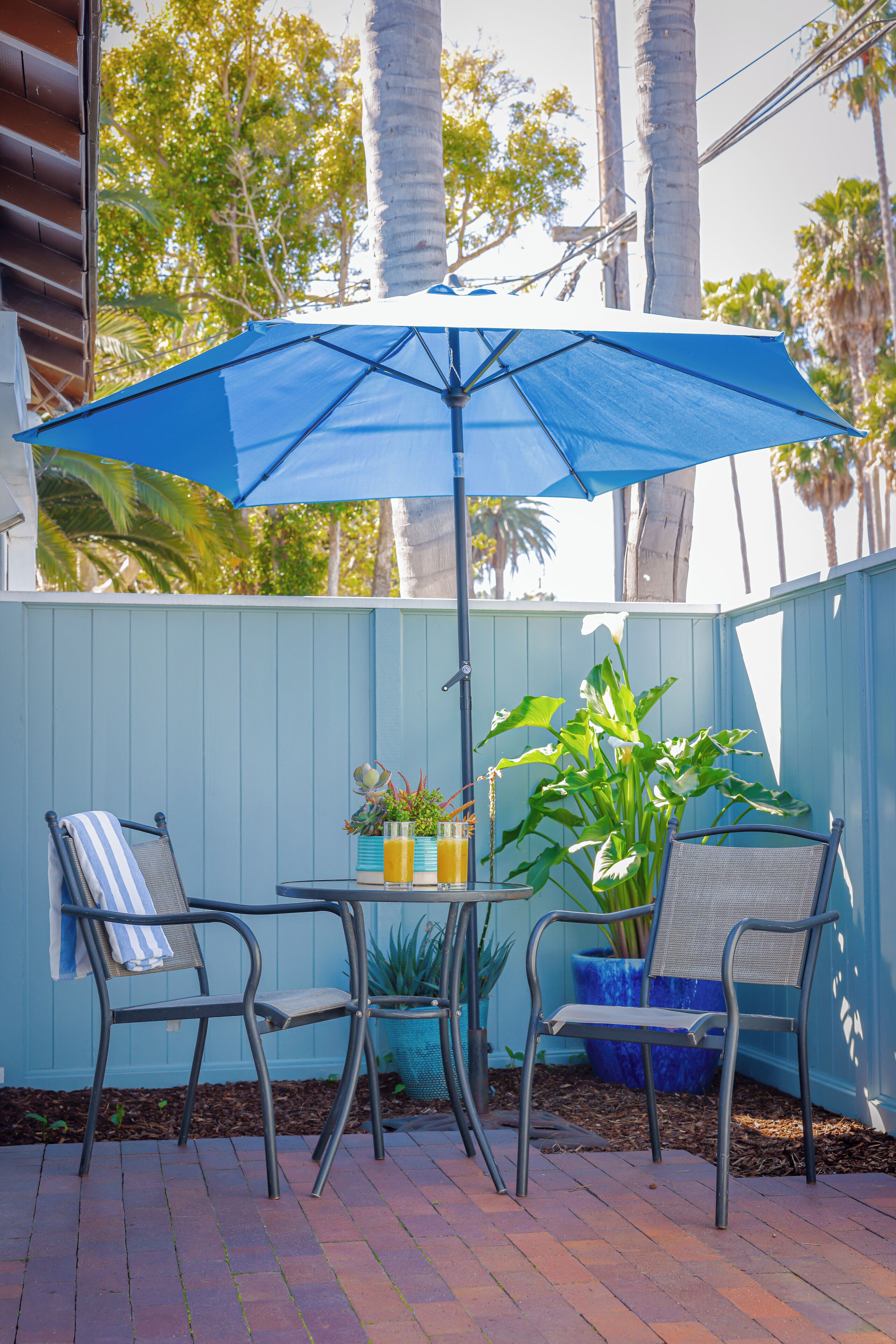 Un patio con dos sillas de metal, una mesa, una sombrilla azul y una planta en maceta.
