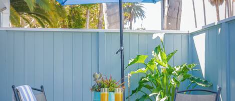 A patio with two metal chairs, a table, a blue umbrella, and a potted plant.