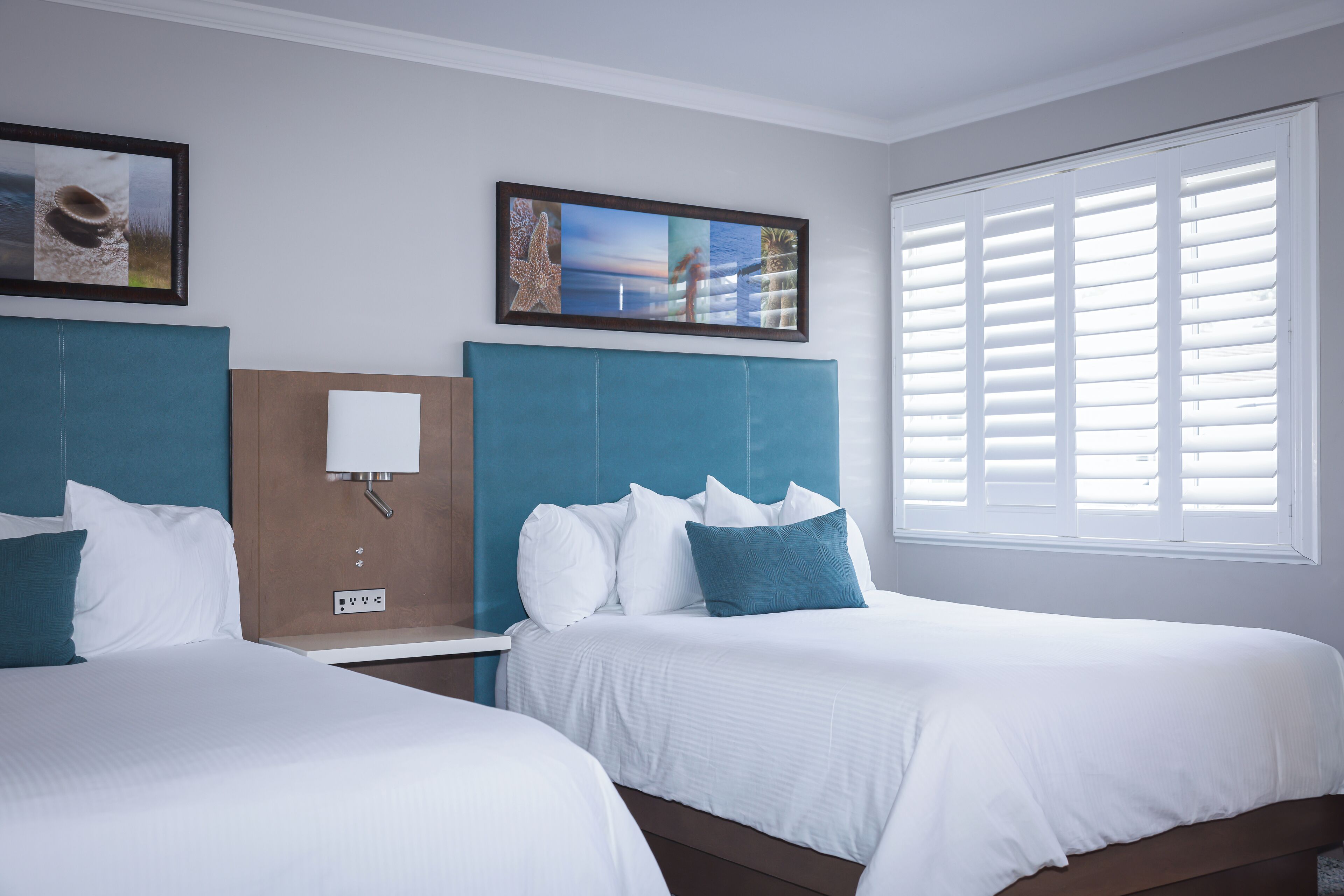 A hotel room with two beds, a large window with blinds, and artwork on the wall.