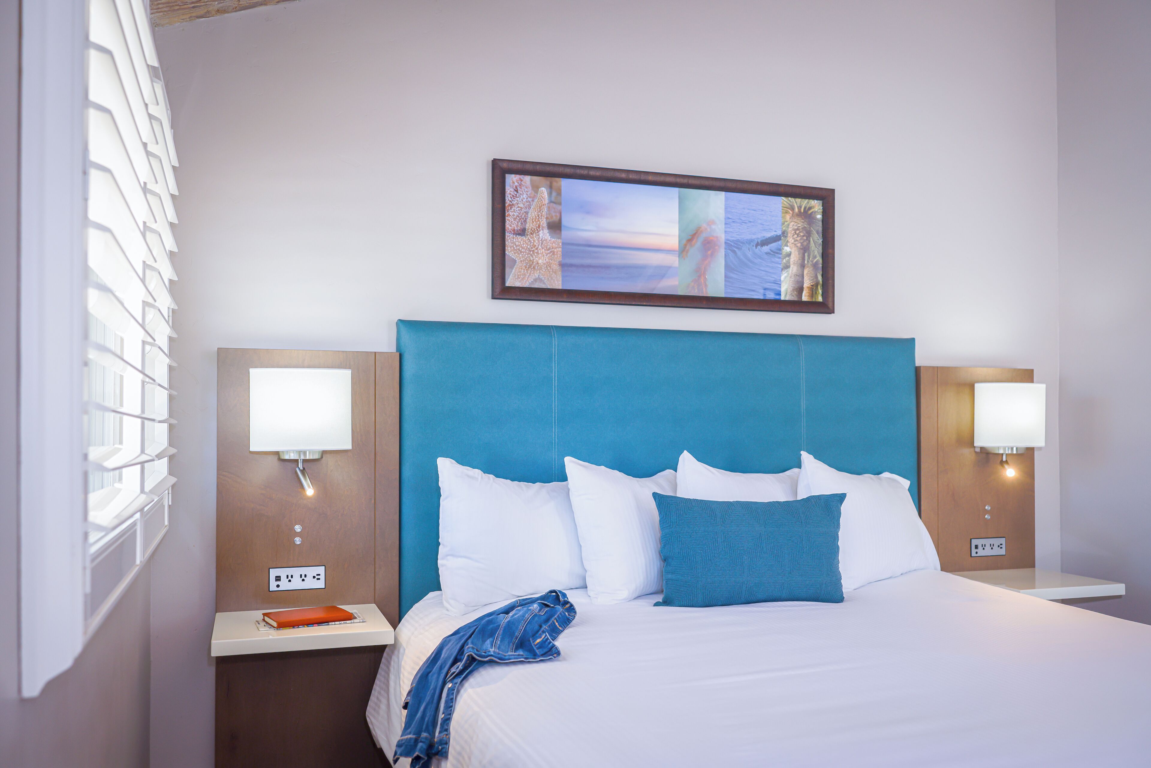A hotel room with a large bed, two bedside tables, a nightstand, a framed picture above the bed, and a window with blinds.