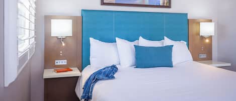 A hotel room with a large bed, two bedside tables, a nightstand, a framed picture above the bed, and a window with blinds.