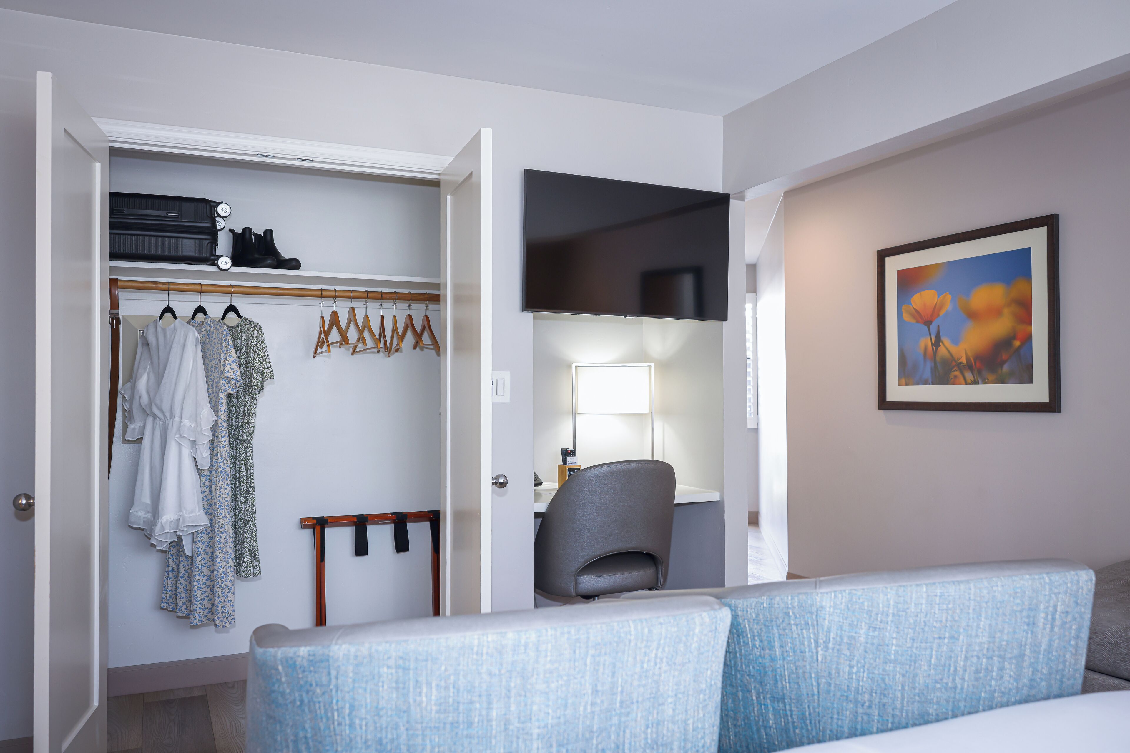 A hotel room with a wardrobe, a desk with a lamp, a grey chair, and a blue couch.