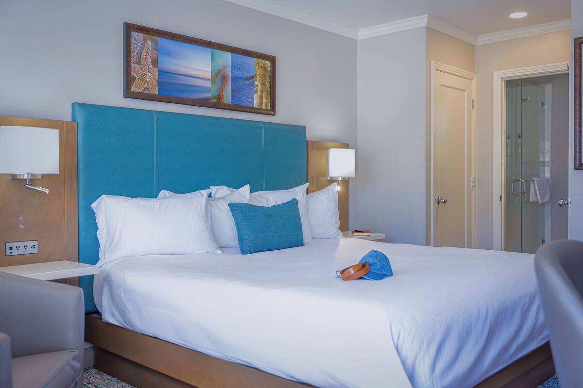 A hotel room with a large bed, a headboard, two bedside tables with lamps, a painting above the bed, and a door to the right.