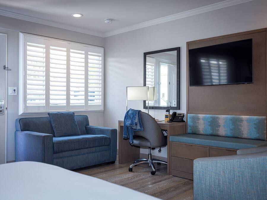 A hotel room with a bed, a desk with a chair, a sofa, a TV mounted on the wall, and a window with blinds.