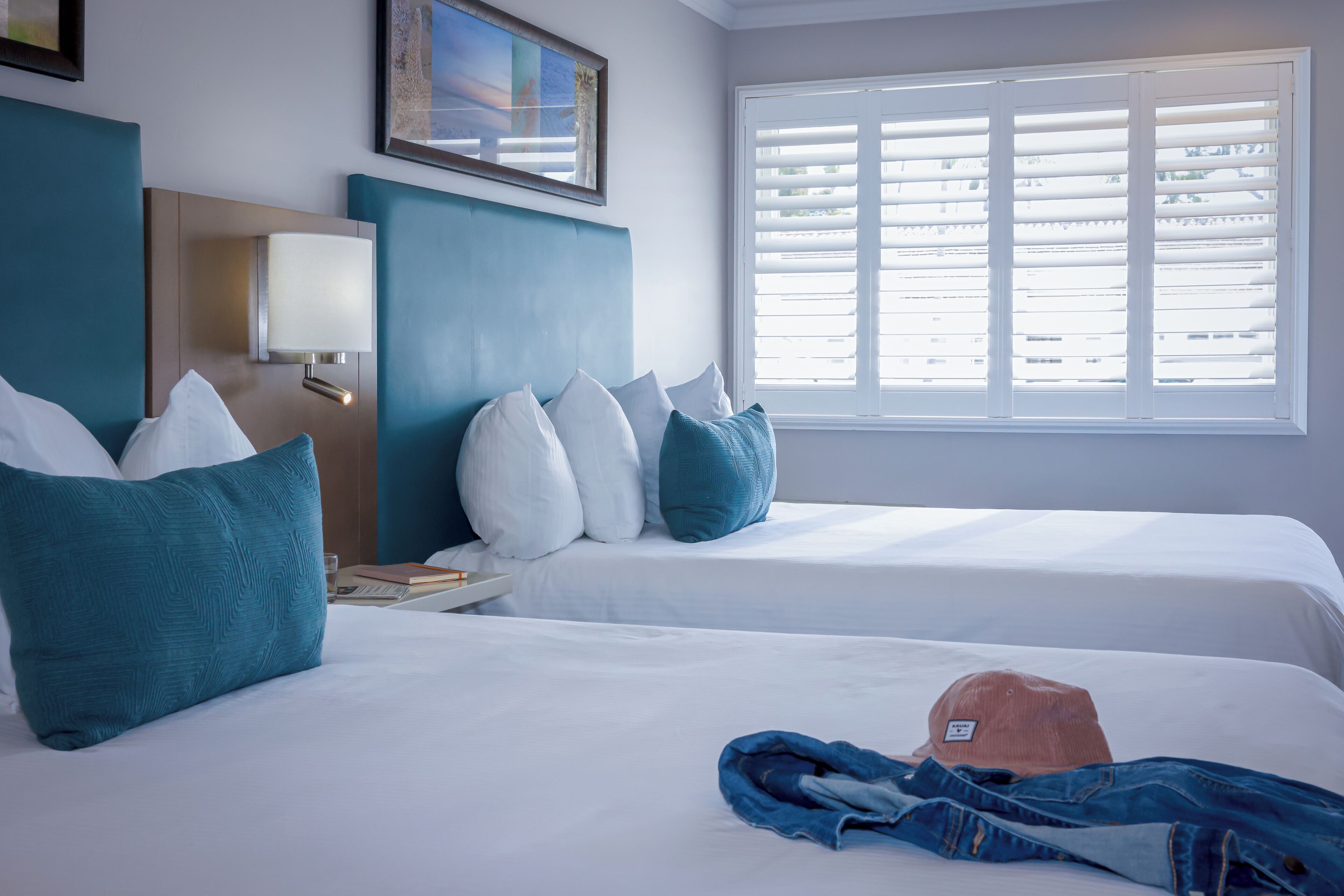 A hotel room with two beds, a nightstand, a lamp, a window with blinds, and a painting on the wall.