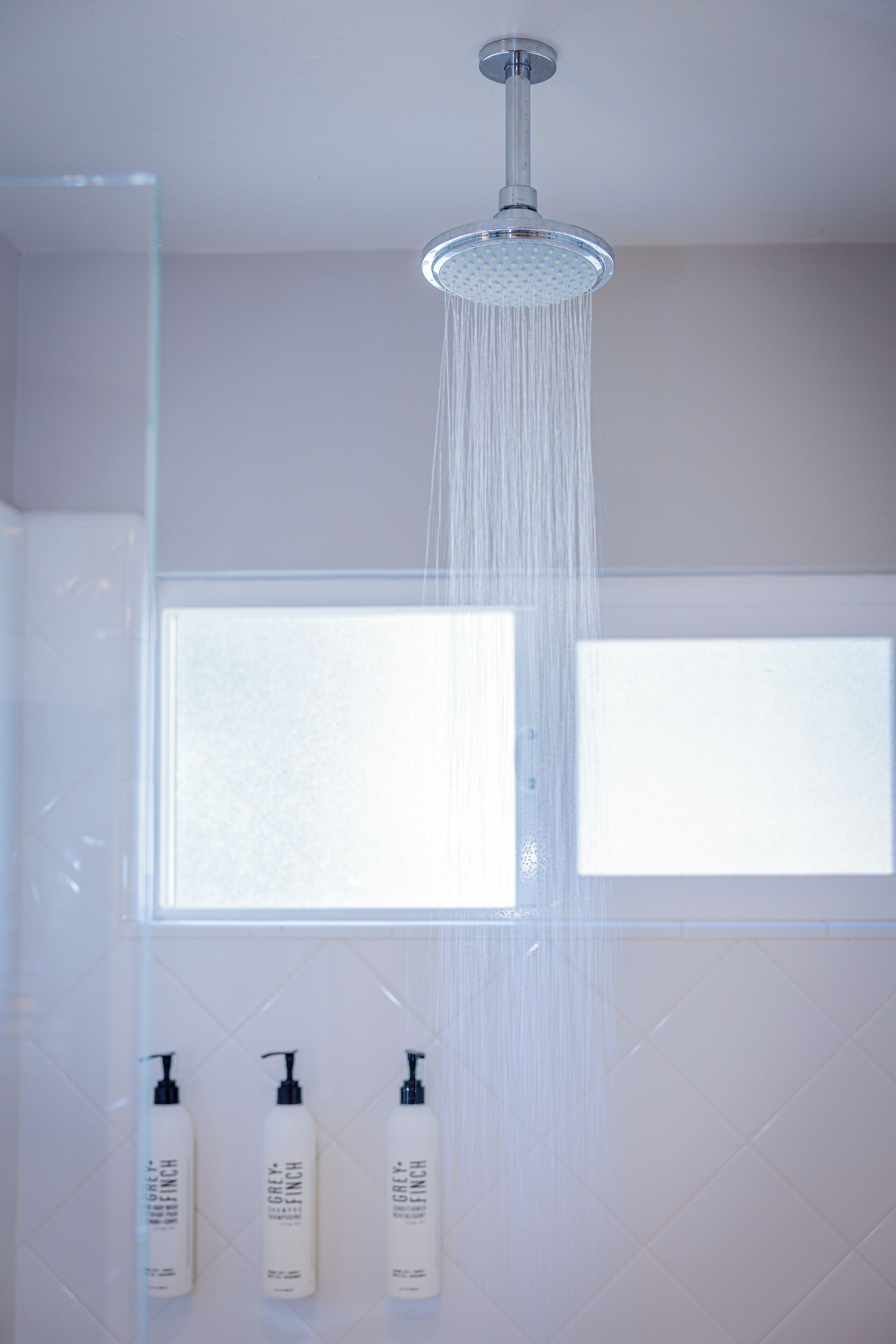A shower with a rain head, a window, and three bottles of Grey Finch shampoo.