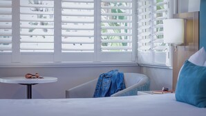 A hotel room with a bed, a round table with a pair of sunglasses and a book, a chair with a denim jacket, and a window with white blinds. - Marina Beach Hotel (Santa Barbara)