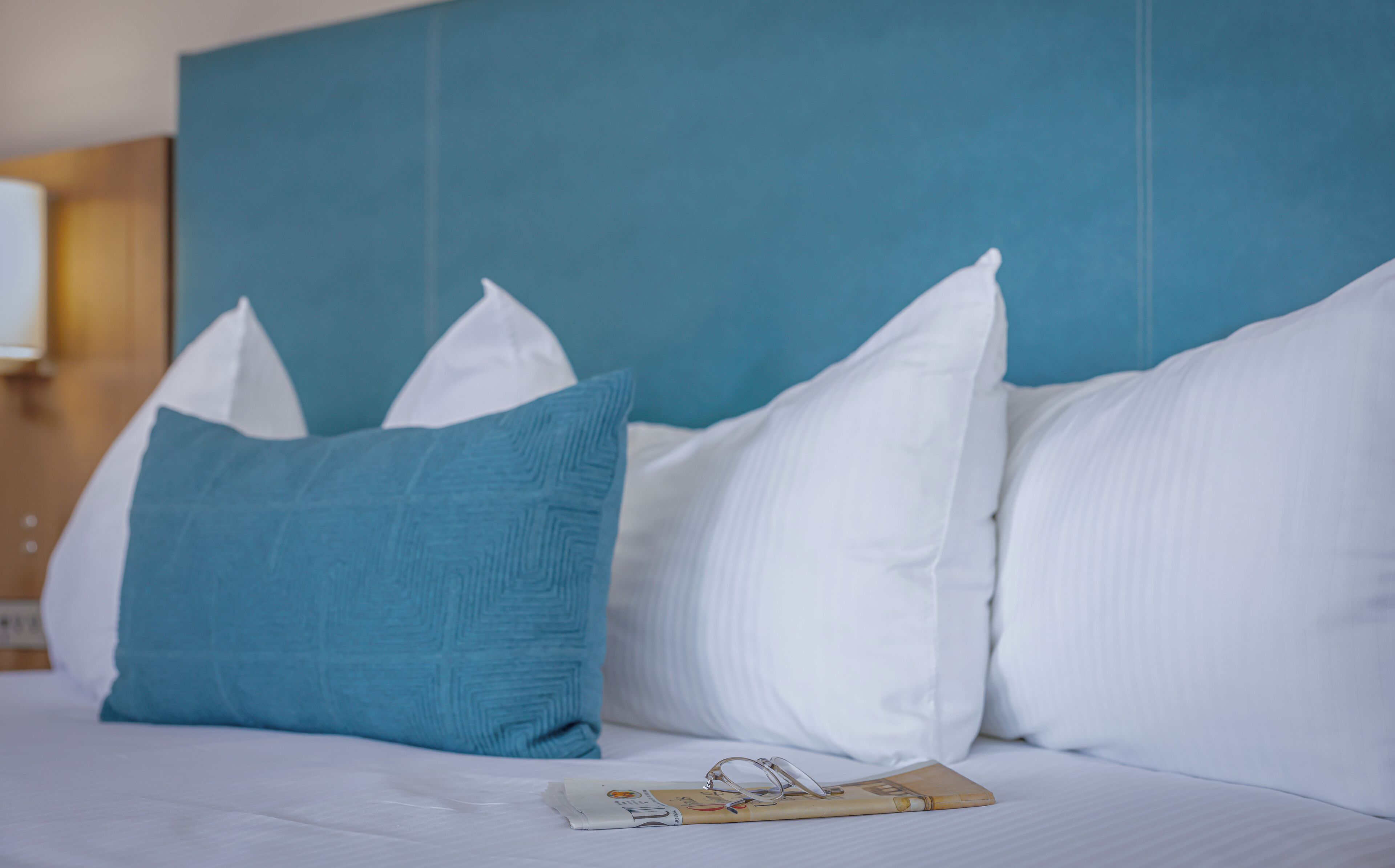 Una cama bien hecha con almohadas blancas y azul turquesa, un par de gafas y una revista sobre la cama.