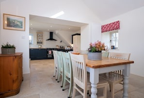 Uma sala de jantar com uma mesa e cadeiras de madeira, cozinha ao fundo e um quadro na parede.