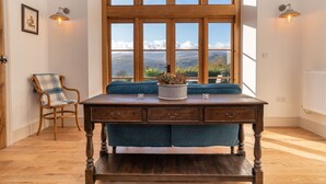 Una mesa de madera con un jarrón de flores, un banco tapizado en azul y una silla con cojín a cuadros en una habitación con ventanas amplias y piso de madera.