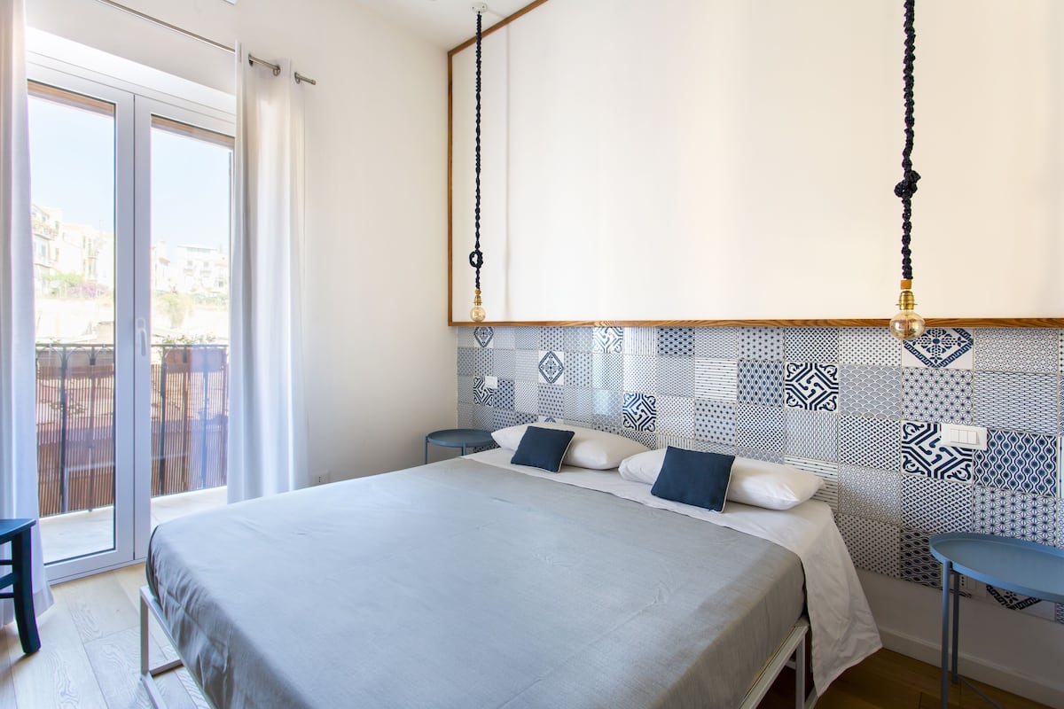 A bedroom with a bed, a small table, a chair, and a view of the outside through a sliding door.