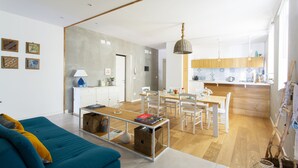 Un salon moderne avec un canapé, un coin repas comprenant une table et des chaises, et une cuisine dotée de meubles en bois et d’un luminaire suspendu.