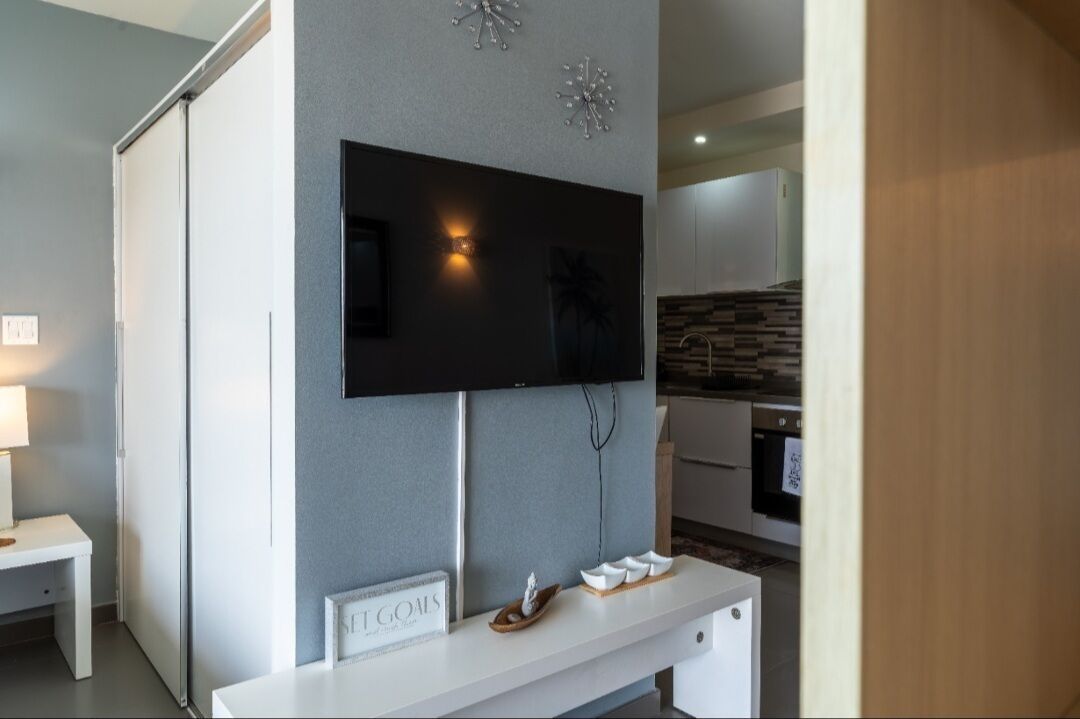 Un salon moderne avec un téléviseur à écran plat fixé à un mur gris, une console blanche sur laquelle est posée une pancarte indiquant « ST GOALS », et un coin cuisine en arrière-plan.