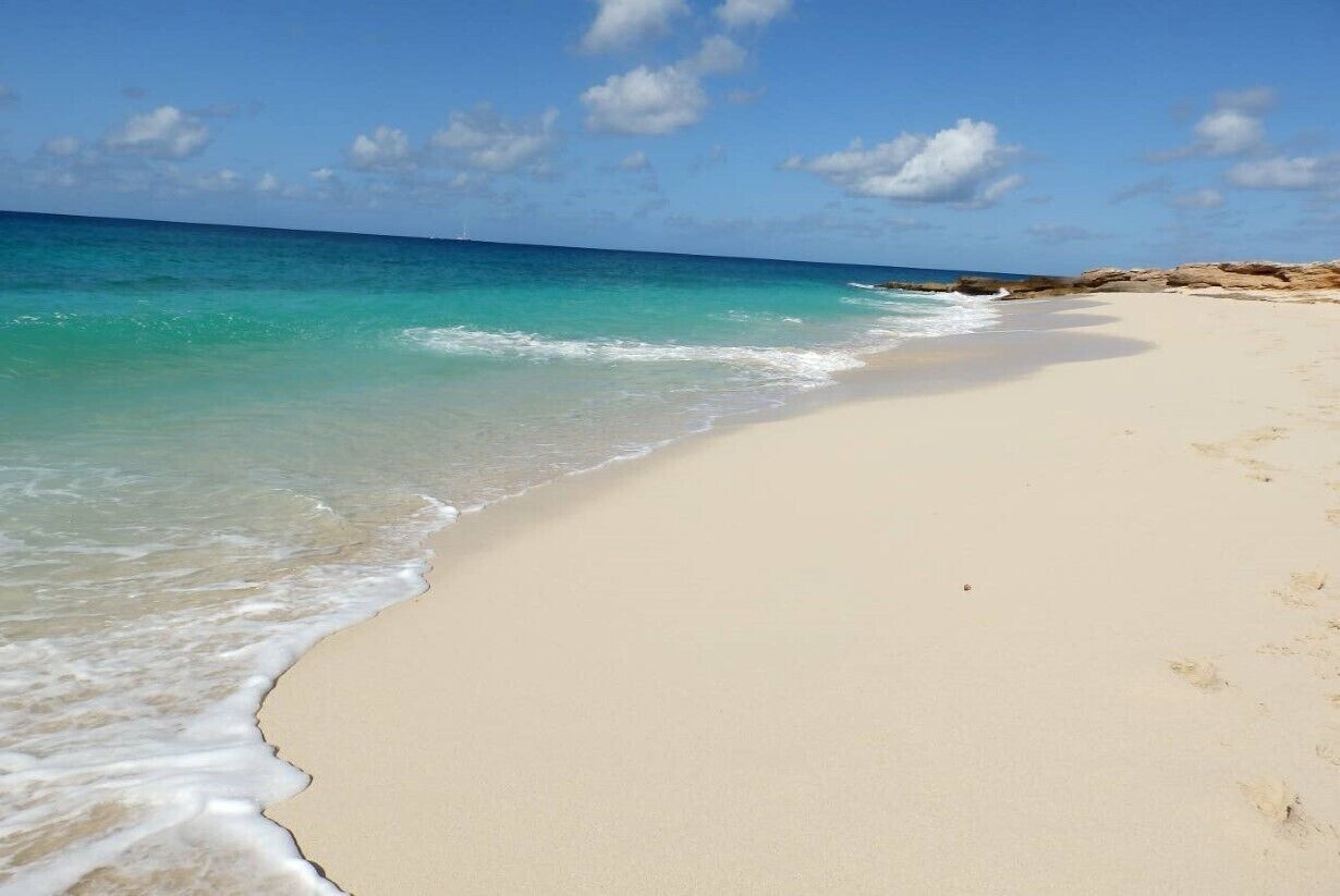 Une plage à l’eau turquoise et limpide, bordée d’un rivage sablonneux et ponctuée de formations rocheuses.