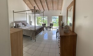 A spacious bedroom with a four-poster bed, a wooden nightstand, a chair, and a large window offering a view of greenery.