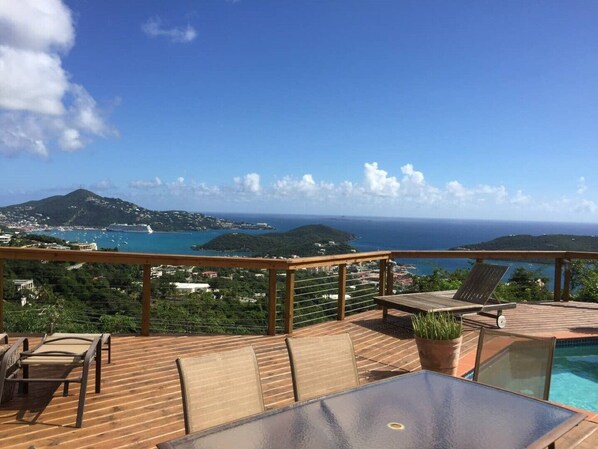 A wooden deck with a glass-top table and chairs, overlooking a coastal view with a clear sky.