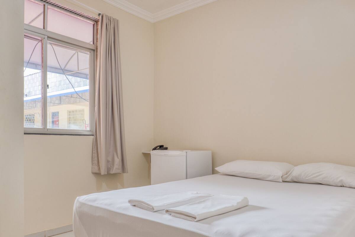 Habitación de hotel con cama, mesita de noche, teléfono y ventana con cortinas.
