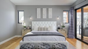 Un dormitorio moderno con cabecera capitonada, una cama grande, un banco y vistas al exterior a través de amplios ventanales.