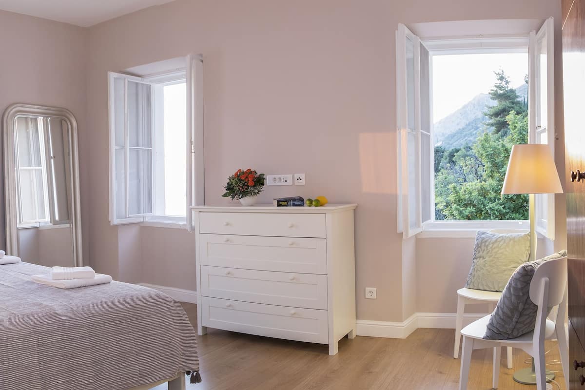 A bedroom with a bed, bedside lamps, a dresser, and a window with a view of trees and mountains.