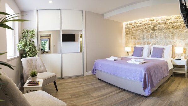Ein modernes Schlafzimmer mit einem großen Bett, einer Wand aus Stein, einer Sitzecke mit einem Sessel und einem kleinen Tisch sowie einem großen Fenster mit Pflanzen.