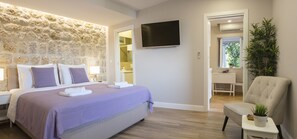 Ein modernes Hotelzimmer mit Wandverkleidung aus Stein, einem Flachbildfernseher, einem bequemen Bett, einer Sitzecke und Blick ins Bad.