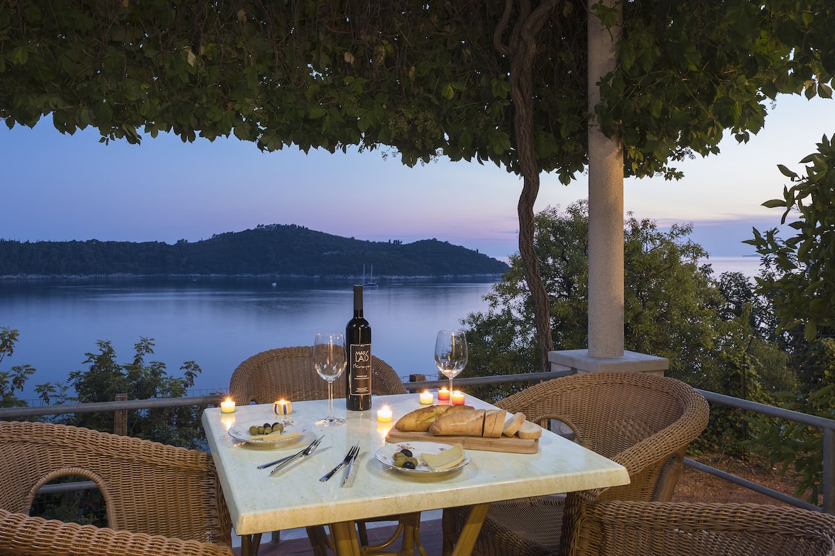 A table set for two with a bottle of wine, glasses, a loaf of bread, and a candle, overlooking a calm lake with a distant mountain.