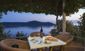 Ein Tisch für zwei, gedeckt mit Wein, Gläsern, Brot und einer Kerze, mit Blick auf einen stillen See und ein fernes Gebirge.