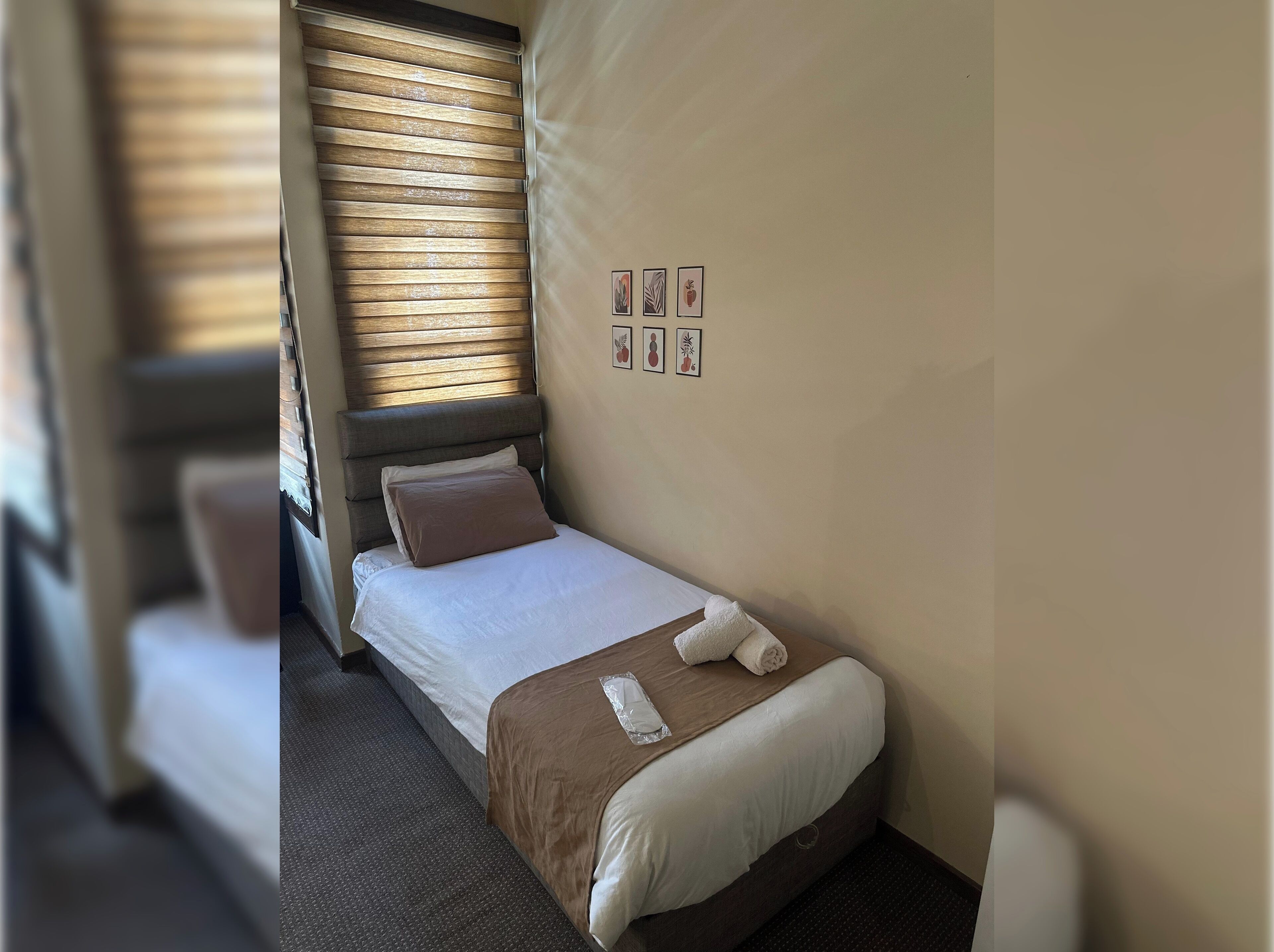 Una cama pequeña y ordenada, con cabecera marrón y ropa de cama blanca, una mesita de noche y una ventana con persianas.
