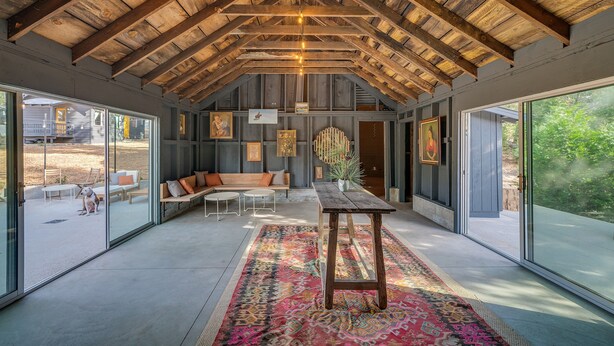 A spacious room with a high ceiling, wooden beams, a large window, a sofa, a table, and a colorful rug.