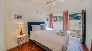 A bedroom with a bed, a ceiling fan, a picture on the wall, and a view of the outside.