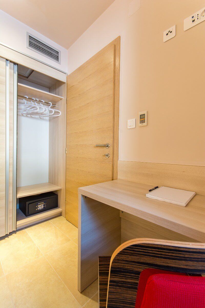 A hotel room with a wooden wardrobe, a desk with a notepad, a red chair, and a wall-mounted air conditioning unit.